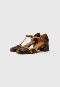 Talons T-strap en cuir marron avec un talon bloc, présentant des rayures horizontales jaunes et marron foncé dans la zone des orteils. Texture lisse.