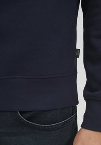 Tricou din bumbac bleumarin cu manșete și tiv cu nervuri, având un etichete mică pe lateral. Asortat cu blugi de denim închis.