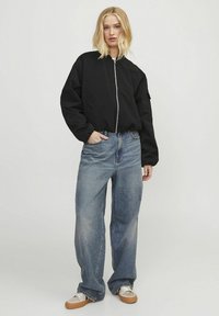 Svart bomberjakke med sølvglidelås, parret med lyseblå vide jeans og brune joggesko. Glatt stoff, avslappet passform, minimalistisk design.