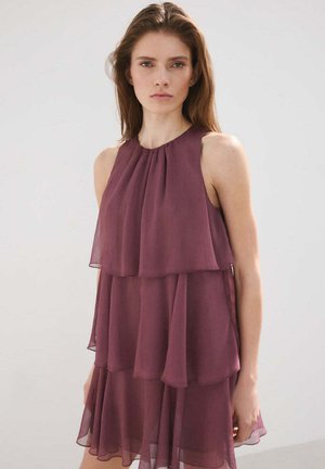 Młoda kobieta o długich włosach, ubrana w warstwową, bezrękawnikową, fioletową sukienkę z szyfonu, stojąca na tle jednolitego jasnego tła.