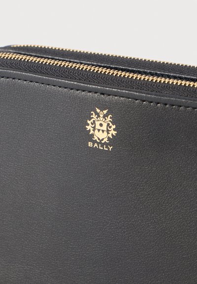 Čierna kožená peňaženka so zlatým zipsom a zlatým logom Bally s erbom a textom na prednej strane.