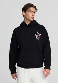 Felpa nera con tasca frontale, caratterizzata da una grafica a forma di stella e testo che recita "CONNECTING" in arancione. Tessuto misto cotone.