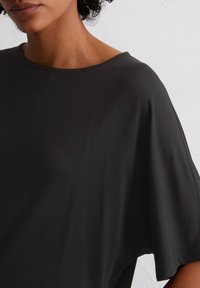 Czarny top z krótkim rękawem z okrągłym dekoltem, wykonany z miękkiego materiału o luźnym kroju, podkreślający gładką fakturę i falbaniasty fason.
