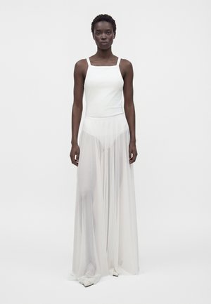 Femme aux cheveux courts portant un haut blanc sans manches et une longue jupe blanche transparente, debout devant un fond blanc uni.