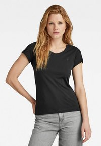 Black cotton t-shirt with a round neckline and short sleeves, featuring a subtle logo detail on the chest, displayed with gray jeans.