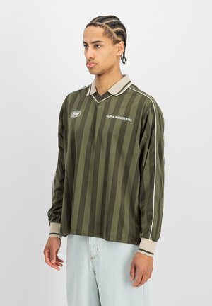 Junger Mann mit geflochtenem Haar, der ein grün gestreiftes langärmeliges Alpha Industries Shirt und hellblaue Jeans trägt, leicht nach links blickend.