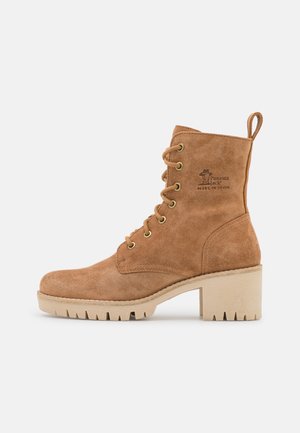 Botte de cheville en suede marron avec un laçage, bout rond, œillets dorés et un talon en caoutchouc épais. Présente un petit logo sur le côté.