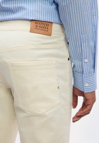 Κοντινό πλάνο σε κρεμ παντελόνι με πίσω τσέπη και δερμάτινο ταμπελάκι του εμπορικού σήματος «Scotch & Soda Amsterdam», φορεμένο με μπλε ριγέ πουκάμισο.