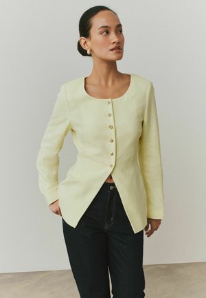 Kvinde iført lys gul knapgjort blazer med en frontsplittelse og mørke bukser, stående med den ene hånd i lommen, og kigger mod højre.