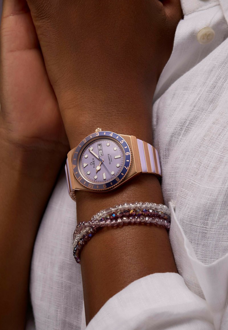 Orologio in oro rosa con lunetta blu e quadrante lavanda, dotato di indici bianchi. Cinturino in silicone a righe lavanda e bianche.