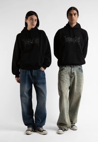 Felpe nere con un design grafico grigio, abbinate a jeans larghi blu e chiari. Entrambi indossano scarpe da ginnastica nere con suole bianche.