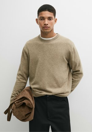 Homme aux cheveux courts portant un pull beige et un pantalon noir, tenant une veste marron pliée dans sa main gauche devant un fond clair uni.