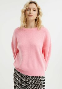 Roze, oversized trui gemaakt van zachte, pluizige stof met een ronde halslijn en verlaagde schouders, gecombineerd met een gestreepte rok.