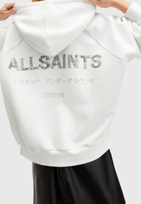 Witte hoodie met een glanzend, zilver geborduurd logo en tekst op de achterkant. Voorzien van een capuchon met trekkoord en geribbelde boorden voor een nauwsluitende pasvorm.