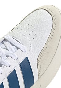 White sneaker with textured leather upper, three navy stripes, suede accent, and white laces. Rounded toe and rubber sole.