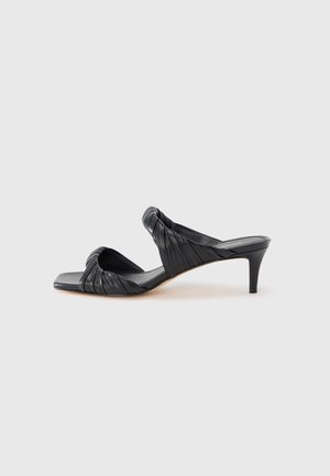 Sandalia mule negra con punta abierta, con tiras de cuero fruncidas y un tacón delgado de altura media sobre un fondo claro y liso.