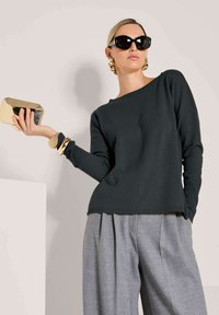 Frau trägt einen schwarzen Pullover und graue weite Hosen, hält eine goldene Clutch, accessoriert mit übergroßen schwarzen Sonnenbrillen und klobigem goldenen Schmuck.