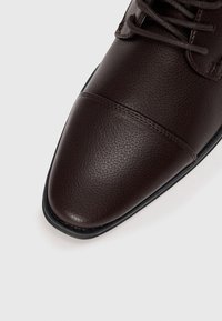 Zapato de vestir de cuero marrón con una superficie texturada, punta afilada y costuras en contraste a lo largo del borde superior y la suela. Diseño con cordones.