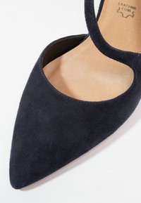 Chaussure plate noire en suède avec bout pointu, semelle intérieure en cuir et design latéral ouvert.