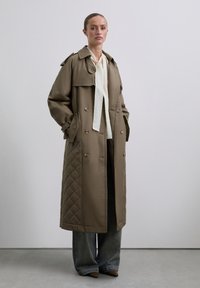 Langer, gesteppter Trenchcoat in Khaki, mit großen Knöpfen, doppelter Knopfleiste, seitlichen Taschen und Schulterklappen. Getragen über einem leichten Oberteil und weiten Jeans.