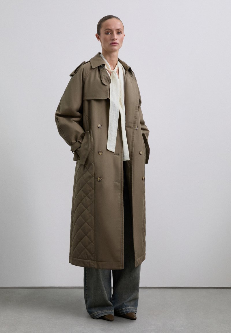 Langer, gesteppter Trenchcoat in Khaki, mit großen Knöpfen, doppelter Knopfleiste, seitlichen Taschen und Schulterklappen. Getragen über einem leichten Oberteil und weiten Jeans.