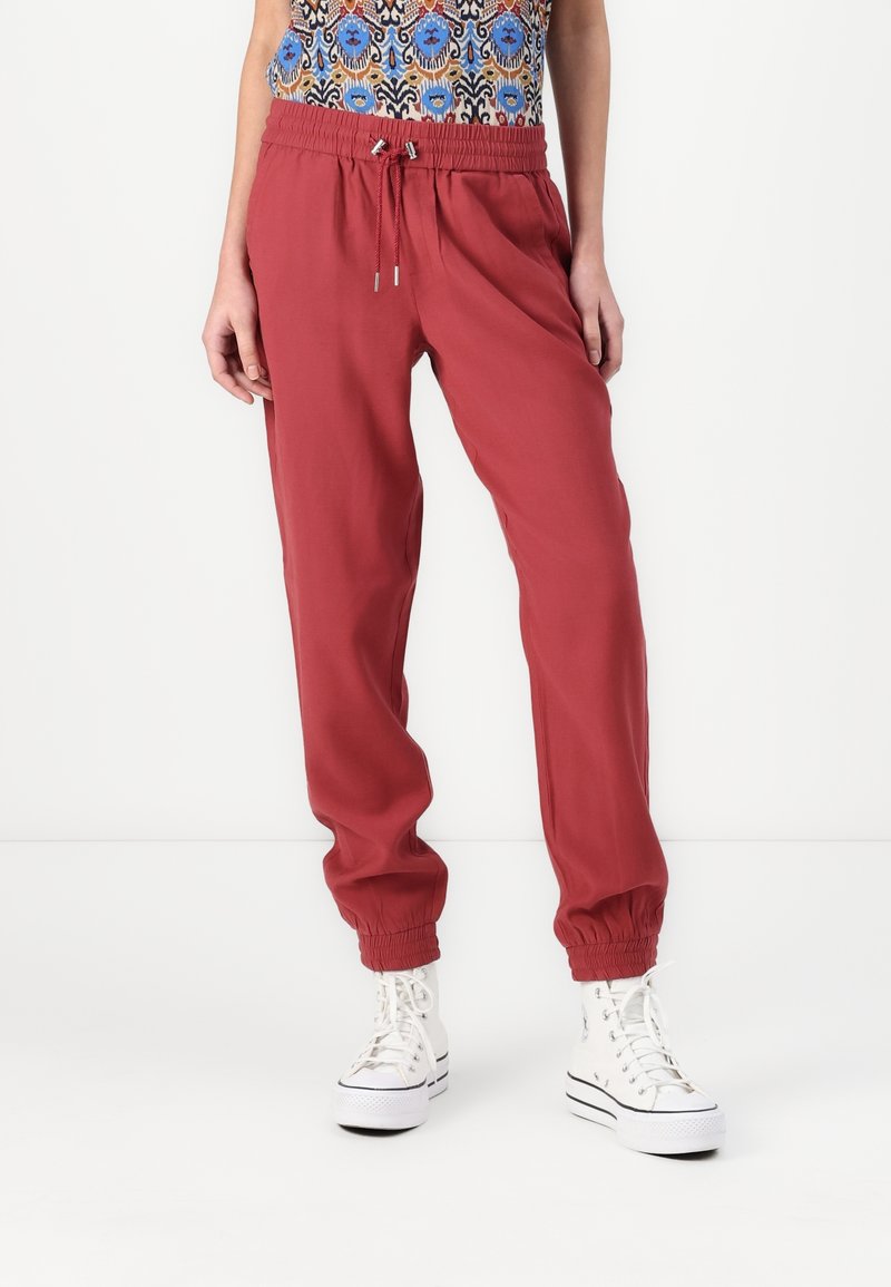 Personne portant un jogger rouge avec une ceinture élastique et des poignets aux chevilles, ainsi que des baskets blanches à plateforme, associée à un haut coloré à motifs.