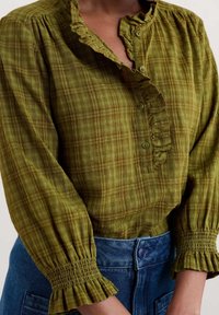 Blouse à carreaux verts avec col à volants et patte de boutonnage. Manches longues avec poignets froncés. Portée avec un short en denim taille haute.