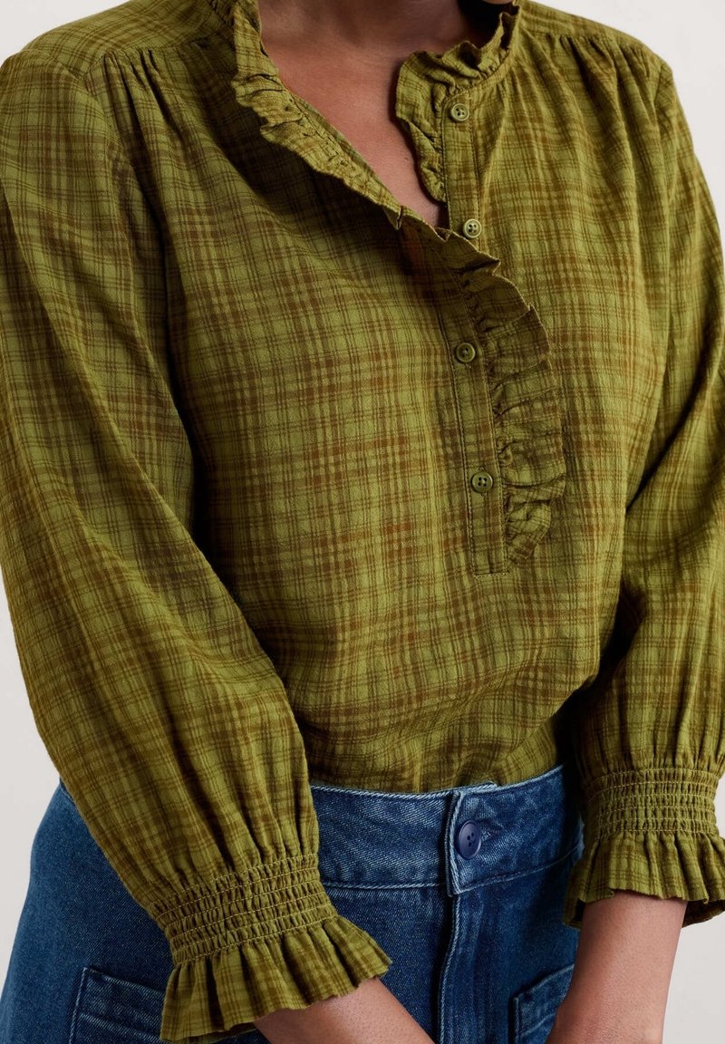 Blouse à carreaux verts avec col à volants et patte de boutonnage. Manches longues avec poignets froncés. Portée avec un short en denim taille haute.
