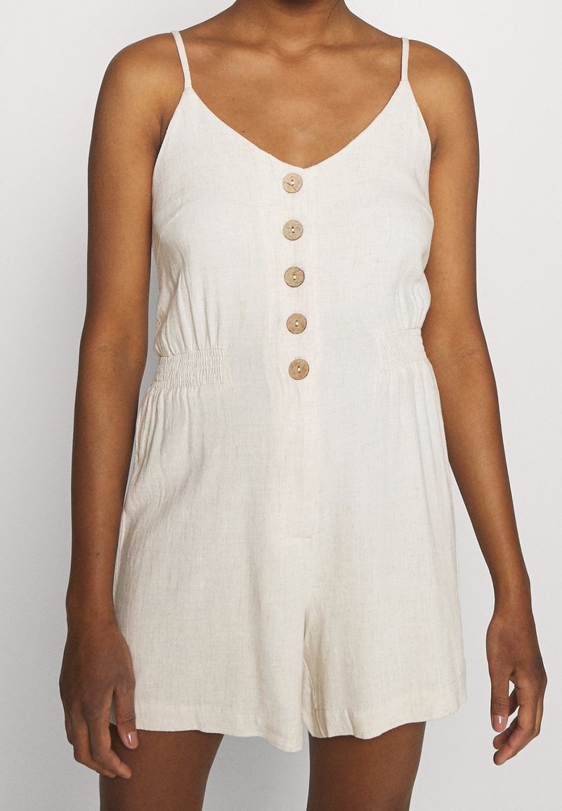 Combinaison en lin de couleur beige clair avec des bretelles spaghetti réglables, un décolleté en V, une fermeture à boutons sur le devant et une taille élastiquée.