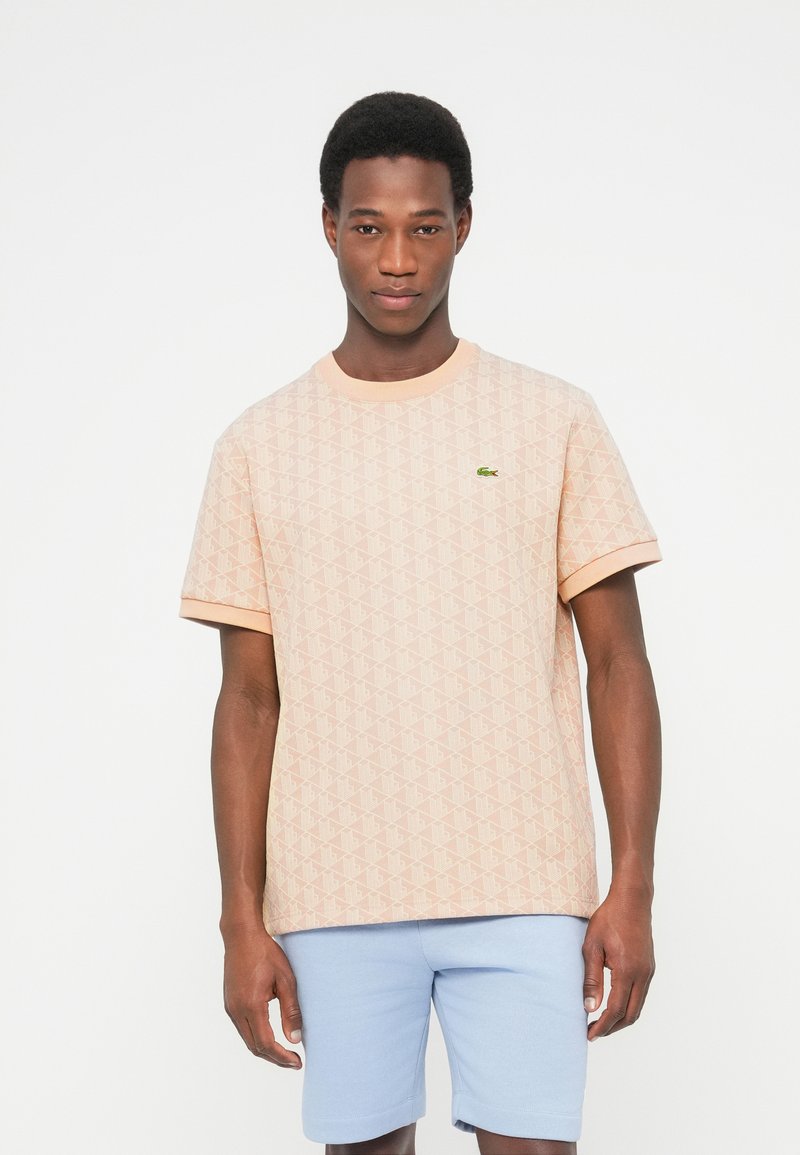 Muž v broskvově vzorované košili s krátkým rukávem se zeleným logem a světlomodrými šortkami stojí před jednobarevným bílým pozadím.