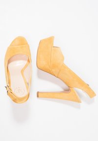 Žluté semišové sandály na vysokém podpatku s odhalenými prsty, nastavitelním popruhem na zadní straně a trojúhelníkovým blokovým podpatkem. Hladký povrch s minimálními detaily.