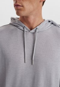 Grå hættejakke lavet af blødt stof, med snøre i halsudskæringen og metal detaljer. Minimalistisk design med diskrete brand detaljer.