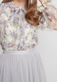 Blouse transparente à imprimé floral avec détails plissés et manches bordées de dentelle, associée à une jupe en maille gris clair ornée de points texturés.