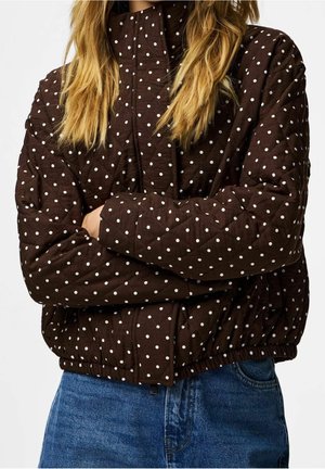 Personne aux longs cheveux blonds portant une veste matelassée marron à pois blancs et un jean bleu, bras croisés.