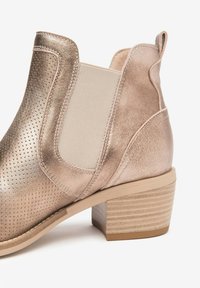 Bottine métallisée rose doré avec surface perforée, panneaux élastiques sur les côtés, petit talon empilé et accents beiges. Fini texturé et design élégant.