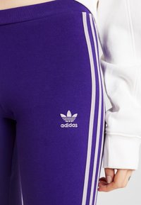 Lila leggings med vita tre-stripes detaljer, texturerad tyg och en broderad Adidas-logga på vänster sida.