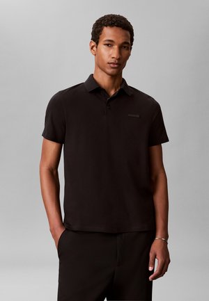 Άνδρας με κοντά σγουρά μαλλιά που φοράει μαύρο polo με κοντό μανίκι και μαύρο παντελόνι, στέκεται με το ένα χέρι στην τσέπη μπροστά σε γκρι φόντο.