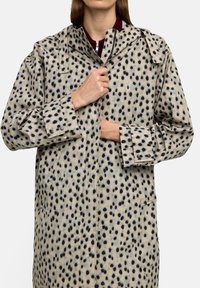 Beżowy płaszcz w czarne kropki; posiada kołnierz, kieszenie z przodu i zapięcia na guzikach. Materiał ma gładką fakturę.