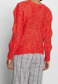 Pull en maille rouge tricoté avec un tissage lâche et texturé, doté de manches longues et d'un ourlet côtelé. Porté sur un pantalon à carreaux gris.