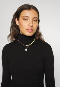 Pull en col roulé noir à côtes, superposé avec une chaîne dorée, un cordon vert fluo et un pendentif floral. Cheveux ondulés naturels et maquillage neutre.