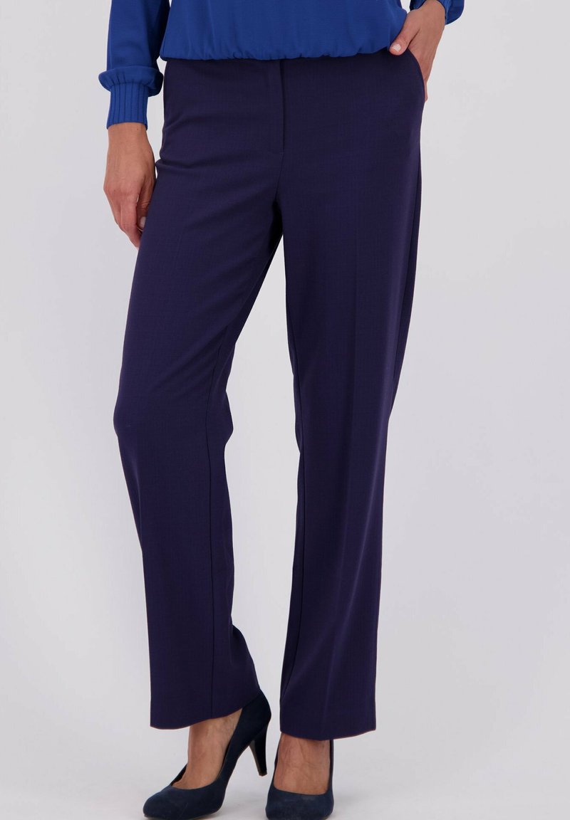 Vrouw in marineblauwe rechte broek met een ingestopte blauwe trui en marineblauwe hoge hakken, staand met één hand in de zak.