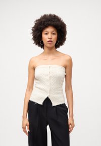 Top strapless de malha creme com padrões de cabos decorativos, apresentando um fecho frontal com botões e uma bainha baguete. Usado com calças pretas.