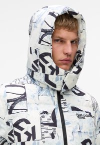 Puchowa kurtka w kolorze białym z niebieskimi i czarnymi wzorami graficznymi. Posiada zamykany zamek z przodu oraz dużą kieszeń. Z logo "KARL LAGERFELD JEANS."