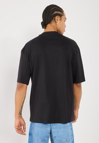 Black oversized t-shirt with short sleeves, round neckline, and a smooth texture. Displayed from the back, with blue denim pants in view.