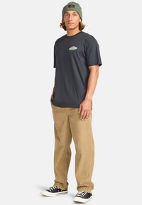 Camiseta de manga corta gris oscuro con un pequeño logo. Combinada con pantalones de pana beige, zapatillas negras y un gorro verde.