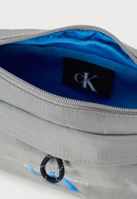 Γκρι υφασμάτινη θήκη με μπλε εσωτερικό, που διαθέτει κλείσιμο με φερμουάρ και μικρή μαύρη θηλιά, καθώς και διακριτικό λογότυπο μπροστά.