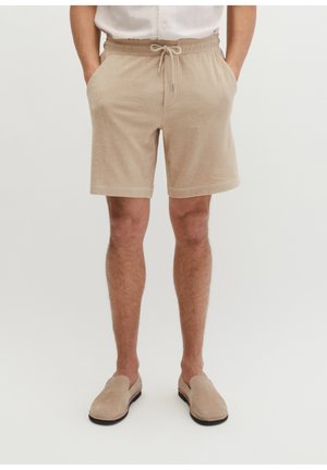 Mężczyzna ubrany w beżowe szorty z frotty z sznurkiem i beżowe buty wsuwane, stojący z rękami w kieszeniach na białym tle.