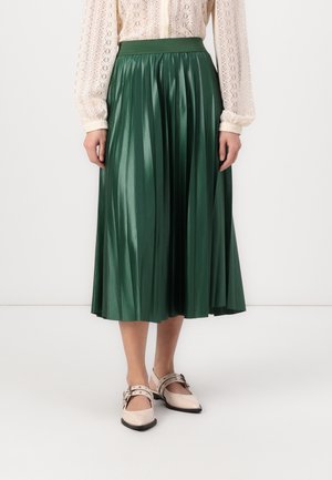 Personne portant une jupe midi plissée vert foncé brillante, un haut en dentelle crème à manches longues et des ballerines beige pointues avec des brides à boucle.
