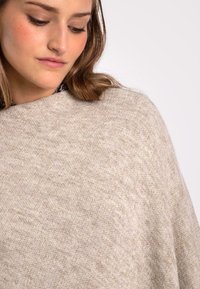 Poncho de punto beige con una textura suave, que presenta un escote amplio y un diseño drapeado. Variaciones sutiles en el color crean profundidad.