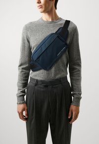 Marine sling bag med en glatt tekstur, med en glidelåslomme og justerbar stropp, brukt diagonalt over en grå genser og stripete bukser.